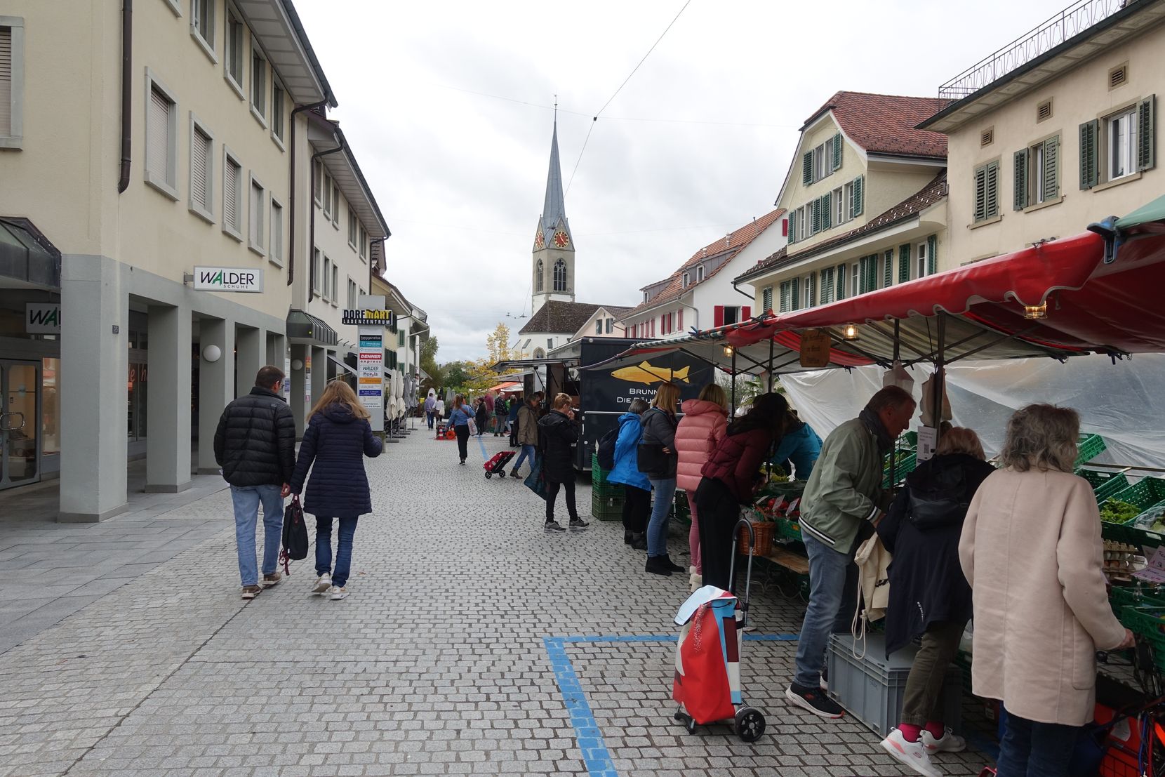 Auf einem gepflasterten Dorfplatz stehen Stand neben Stand, wo Leute durchschlendern.